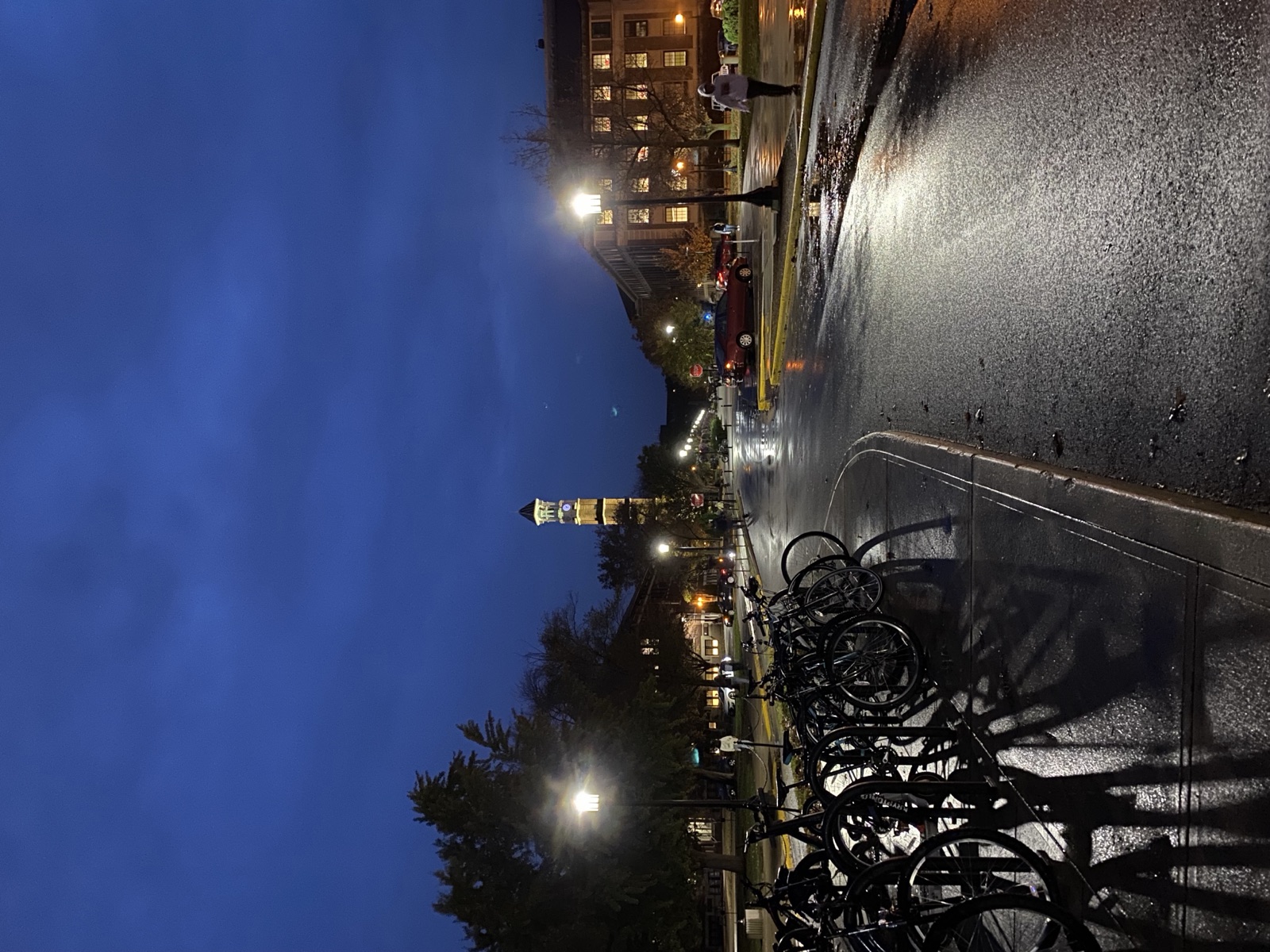 The Purdue University bell tower lit up at dusk at the end of a rain-slicked campus street, with a long row of parked bikes in the foreground.