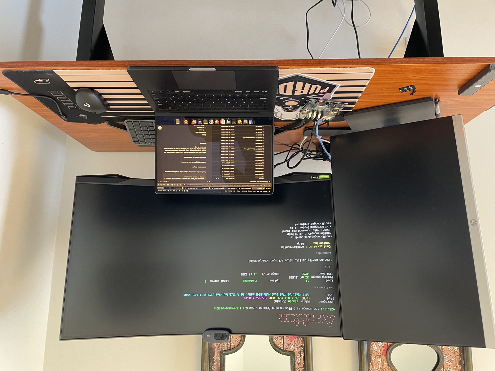 A home desk setup: an ultrawide monitor showing an Armbian login banner for an Orange Pi 5 Plus, the single-board computer itself wired up on a Purdue desk mat, and a MacBook open beside it.