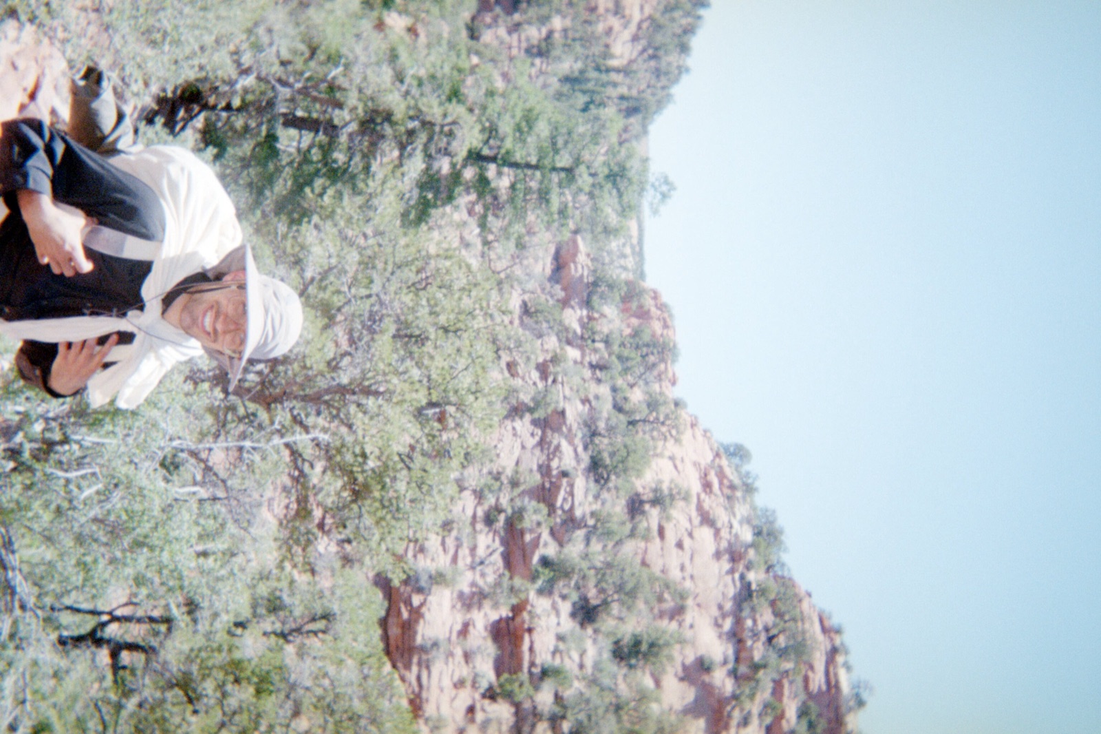 Film photo of a hiker in a wide-brimmed sun hat grinning on a trail below red sandstone canyon walls dotted with juniper.