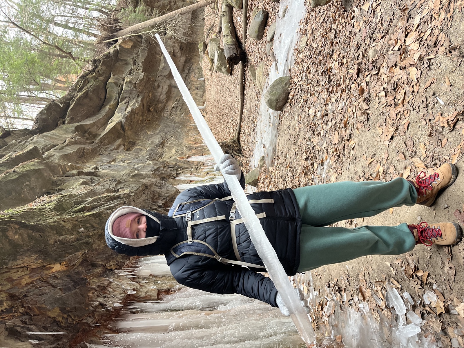 A bundled-up hiker holding a giant icicle horizontally like a spear, in front of a sandstone canyon wall hung with frozen waterfalls.