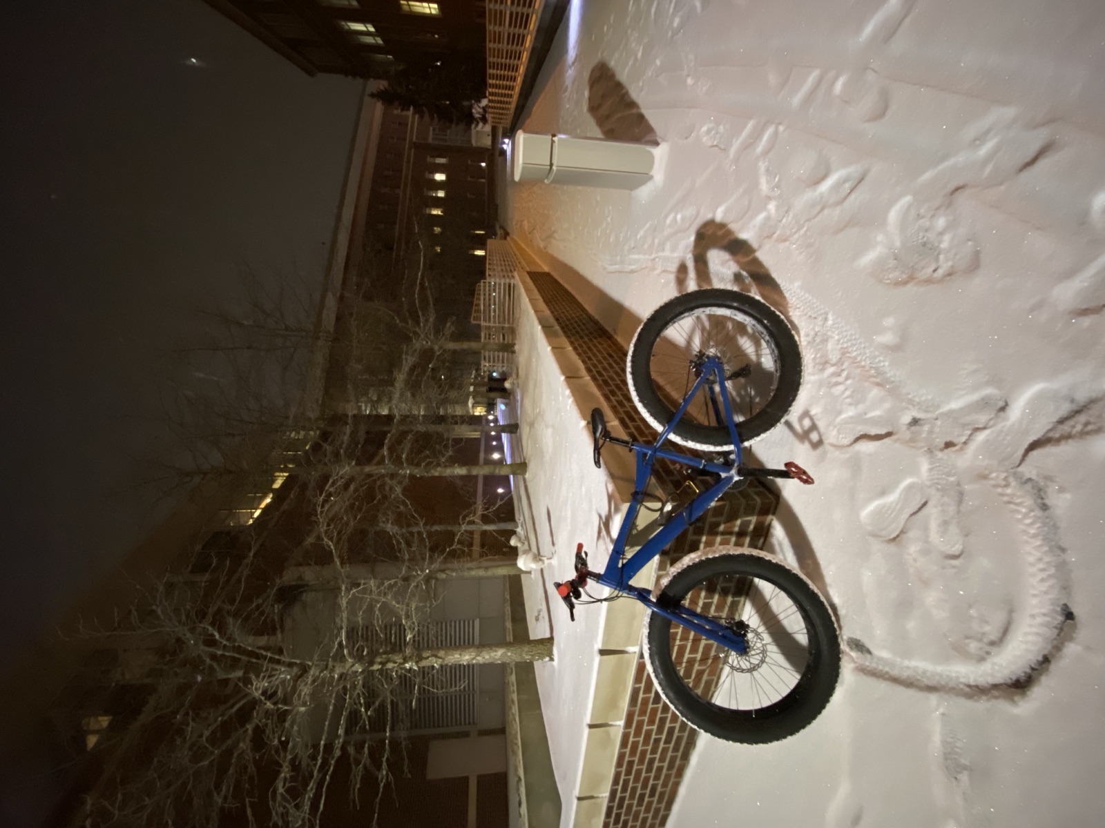 The same blue fat-tire bike at night on a snow-covered Purdue campus walkway, its tire tracks curving away through fresh powder.