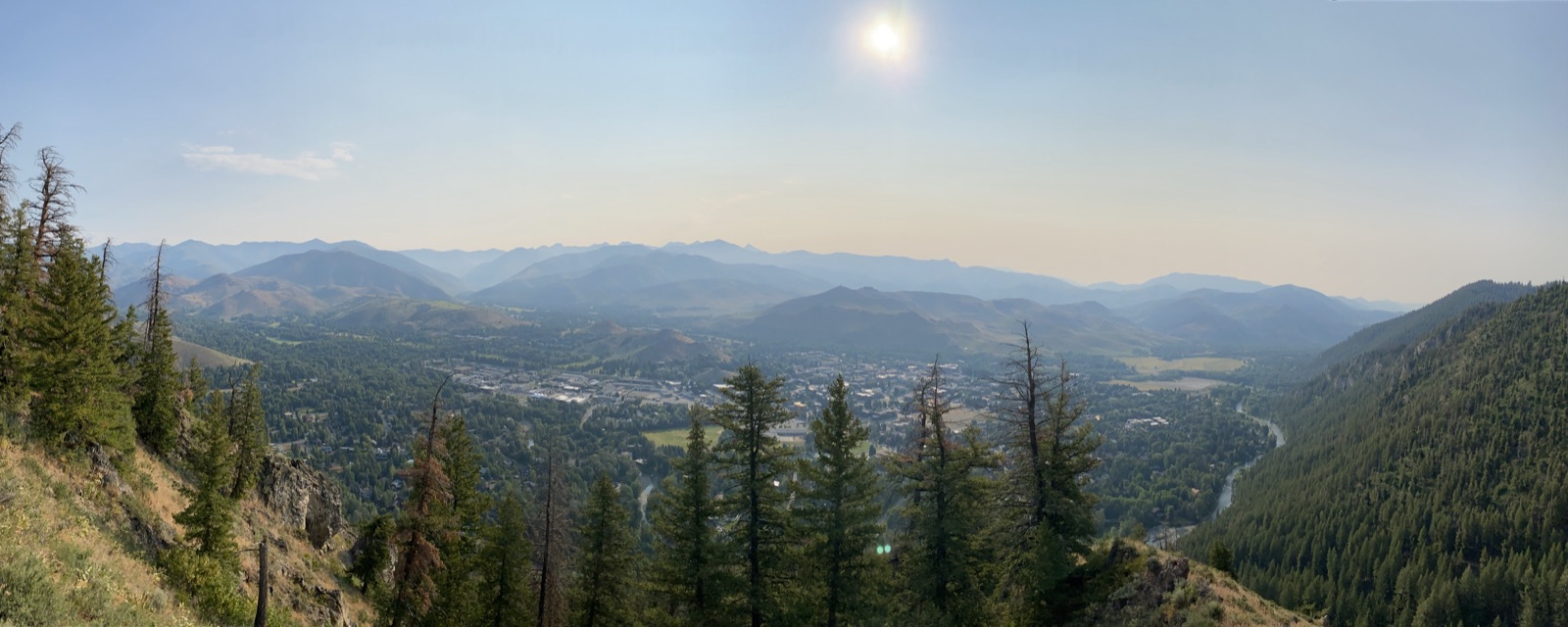 Panorama from a pine-covered ridge looking down over a mountain town in a wide green valley, with hazy blue peaks stacked to the horizon.